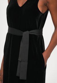 Vestido negro sin mangas en suave terciopelo con un escote en V profundo y un lazo en la cintura negro liso. Cuenta con bolsillos laterales y una silueta elegante.