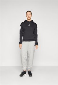 Sweatshirt preto com capuz e riscas brancas nas mangas e no bolso frontal, combinado com calças de treino cinzas e sapatilhas de desporto pretas.