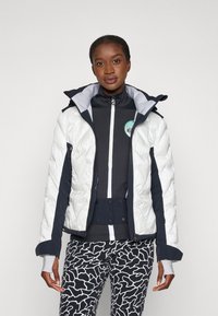 Chaqueta blanca acolchada con detalles en azul marino, bolsillos con cremallera y capucha. Lleva puesta sobre una blusa oscura y pantalones de rayas en blanco y negro.