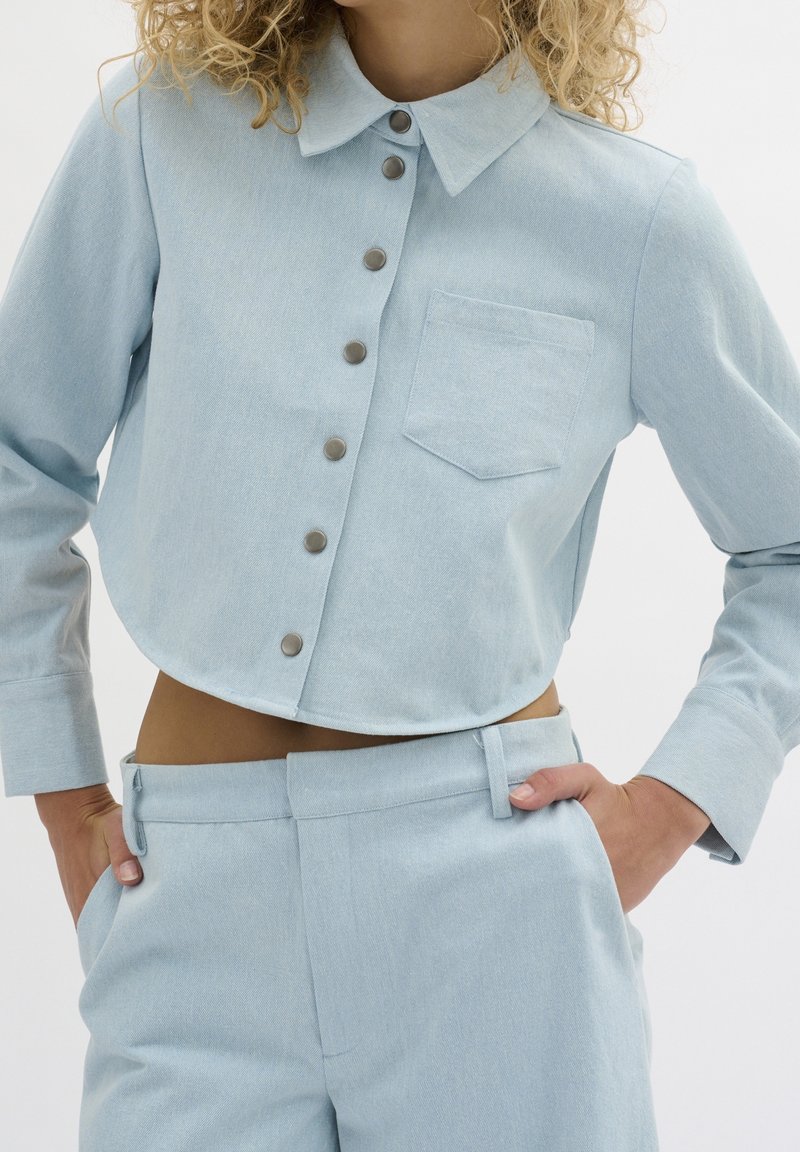 Chemise courte bleu clair en tissu texturé avec col, poche avant et cinq boutons en métal. Associée à un pantalon assorti.