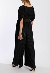 Schwarzer plissierter Overall mit Puffärmeln, kontrastierendem grünen Leopardenprint-Gürtel und weitem Bein. Schwarze Clutch in der Hand.