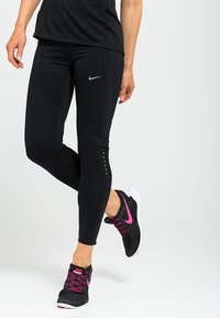Black leggings featuring a discreet Nike logo, textured accents, and reflective dots near the hem, paired with black and pink athletic shoes.