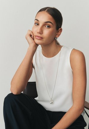 Young woman with tied-back hair wearing a white sleeveless top, silver pendant necklace, and black pants, resting her head on her hand.