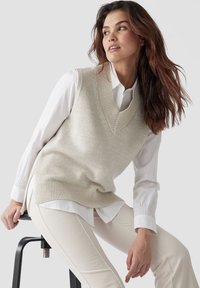 Beige gestrickte Weste mit V-Ausschnitt über einem weißen Hemd mit Knöpfen, kombiniert mit cremefarbenen, enganliegenden Hosen. Gleiche Textur, leichtes Material.