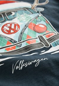 Grafisk T-shirt med en Volkswagen transporter med et ferietema, farverige geometriske mønstre og hvid tekst på en mørk stofbaggrund.