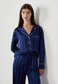 Marinblå satinpajamaset med vita vertikala ränder, som har en knäppningstopp med ficka och byxor med elastisk midja. Mjuk textur.