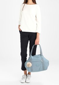 Sac fourre-tout bleu avec bretelles doubles, une poche avant zippée et un détail pompon duveteux ; tissu lisse ; assorti à un pantalon rayé et des chaussures blanches.