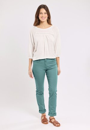 Femme souriante portant un chemisier blanc à manches trois-quarts, un pantalon slim bleu sarcelle retroussé aux poignets, et des sandales en cuir marron, debout sur un fond uni.