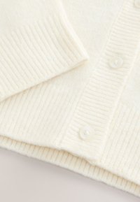 Krēmkrāsas kardigans no mīksta rievota auduma, ar pogājamu priekšpusi, kurā ir apaļas baltas pogas un ar teksturētām aprocēm.