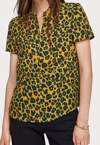 T-shirt à manches courtes de couleur jaune vif avec un imprimé léopard noir et vert, présentant un col en V et des coutures en pointillé le long des seams.