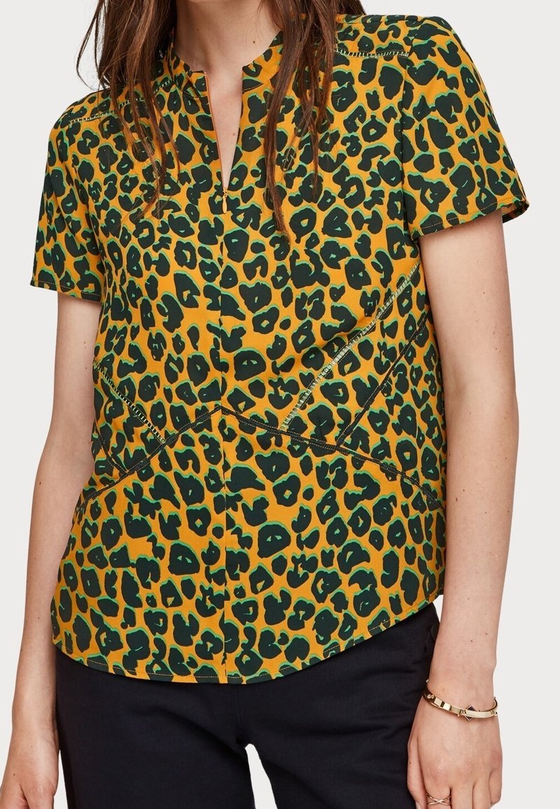 T-shirt à manches courtes de couleur jaune vif avec un imprimé léopard noir et vert, présentant un col en V et des coutures en pointillé le long des seams.