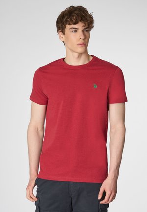 Un uomo con i capelli corti castani indossa una maglietta rossa con un logo verde, in piedi contro uno sfondo grigio chiaro. Guarda leggermente di lato.
