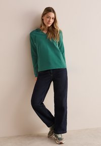 Grön huvtröja med V-ringning och dragsnören, kombinerad med mörkblå vida jeans och gröna och vita sneakers.