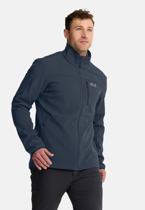 Uomo con capelli ricci che indossa una giacca softshell blu navy e pantaloni neri, posato mentre cammina su uno sfondo bianco.