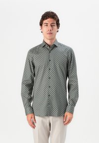 Mann, der ein dunkelgrünes Button-Up-Hemd mit weißen kreisförmigen Mustern und beigen Hosen trägt und vor einem schlichten weißen Hintergrund steht.