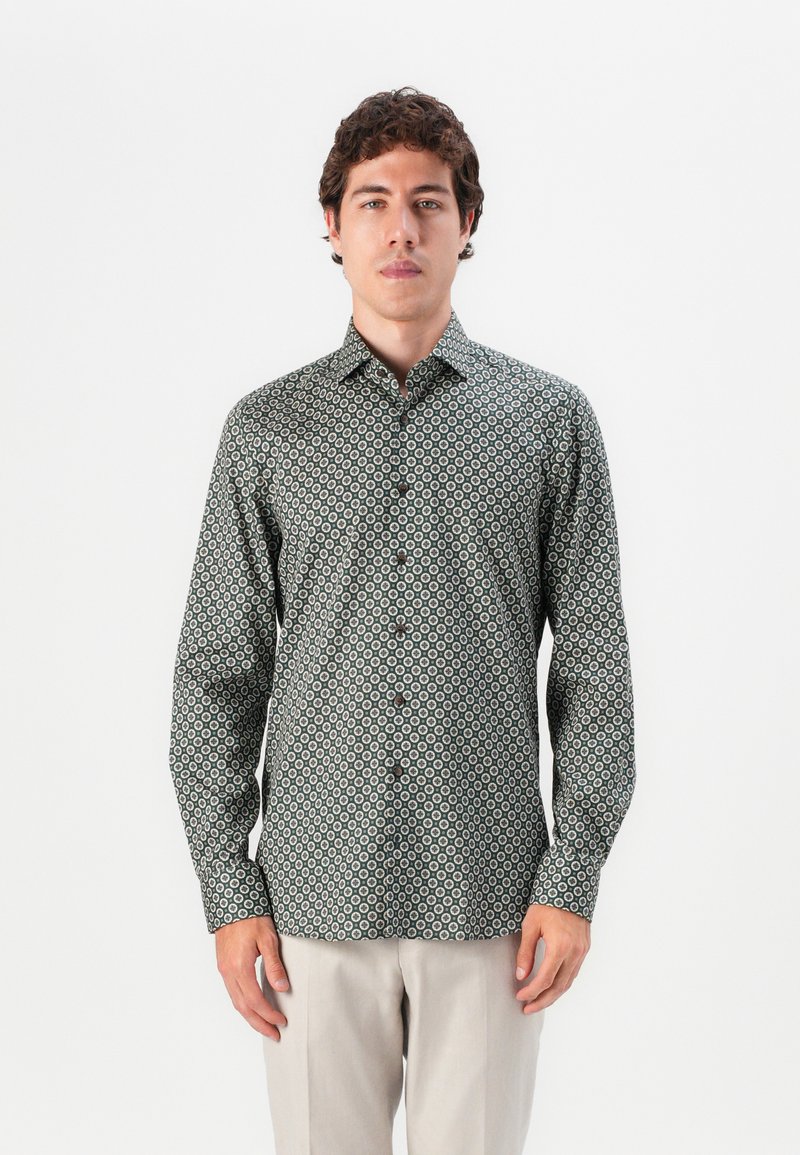 Mann, der ein dunkelgrünes Button-Up-Hemd mit weißen kreisförmigen Mustern und beigen Hosen trägt und vor einem schlichten weißen Hintergrund steht.