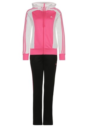 Kobieta w różowej bluzie z kapturem, pasującej różowej koszulce, czarnych spodniach dresowych z różowymi paskami oraz jasnoniebieskich tenisówkach, stojąca na szarym tle.