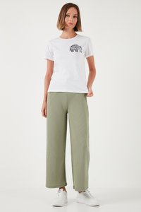 Frau trägt weißes T-Shirt mit schwarz gemustertem Elefantenmotiv, hellgrüne weite Hosen und weiße Turnschuhe, steht vor weißem Hintergrund.