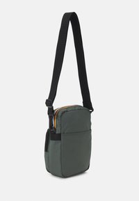Zelená crossbody taška s polstrovaním a čiernym popruhom, so zipsovým uzáverom a kontrastnými zipsovými akcentmi. Obsahuje jednu prednú kapsu pre dodatočné úložisko.
