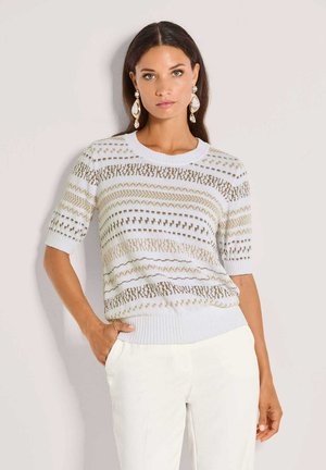 Vrouw met lang donker haar draagt een wit gebreid topje met beige patronen, witte broek en grote hangende oorbellen, staand met één hand in de zak.