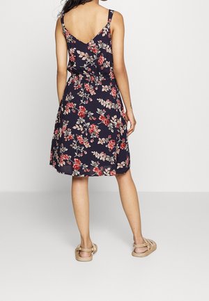 Femme portant une robe sans manches bleu marine avec un imprimé floral rouge et rose, debout pieds nus sur un fond blanc uni.