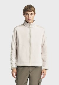 Helle beige Zip-up-Jacke mit hohem Kragen, strukturiertem Stoff und zwei Fronttaschen. Glatte Nähte und subtile Akzente an den Schultern.
