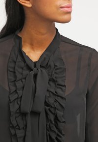 Blouse noire transparente avec un design à volants sur le devant et un col noué. Le tissu est lisse avec une texture légère.