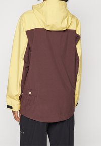 Färgglad jacka med brun nedre del och gul övre del, med huva och en sidoficka.