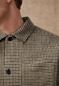 Giacca a motivo houndstooth marrone e beige con colletto a punta, bottoni neri e tasca sul petto con tessuto testurizzato.