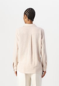 Light beige long-sleeve shirt, textured fabric, collar, back pleat detail, buttoned cuffs, relaxed fit, worn with white trousers.