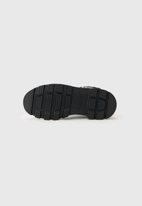 Suela de zapato negra con un diseño texturizado y robusto, con ranuras profundas para mayor tracción. El material parece ser goma y se puede ver la marca.