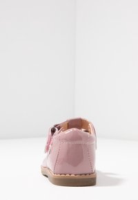 Różowe lakierowane buty z zaokrąglonym noskiem, otwartym obcasem i pojedynczym paskiem na rzep. Posiadają beżową gumową podeszwę oraz ozdobne przeszycia.
