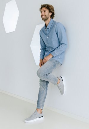 Sneakers in suede grigio con suole bianche e dettagli neri, abbinati a una camicia di jeans blu chiaro e jeans sbiaditi, davanti a un muro neutro.