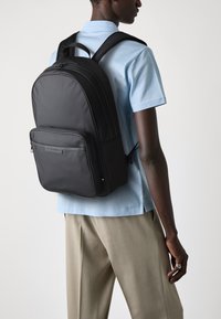 Black backpack made of nylon with two front zippers, adjustable straps, and a smooth finish. Worn on a blue shirt and beige trousers.