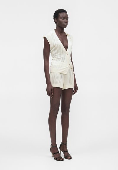 Γυναίκα που φοράει αμάνικο εκρού πλεκτό γιλέκο με κουμπιά και ταιριαστό σορτς, σε συνδυασμό με μαύρα σανδάλια με τακούνι, στέκεται μπροστά σε λευκό φόντο.
