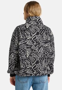 Svart-hvitt mønstret fleecejakke med høy krage, oversized passform og ribbede mansjetter. Har abstrakte geometriske design.