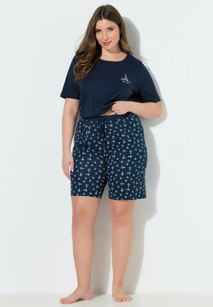 Kvinde med langt brunt hår iført en mørkeblå t-shirt og mørkeblå shorts med hvide ankerprint, stående barfodet mod en hvid væg.