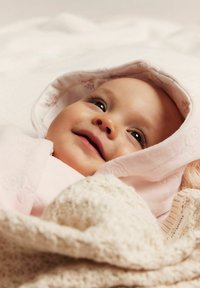 Lichtroze babydeken met bloemenkap, zachte textuur. Het gezicht van een baby met donkere ogen en een lichte glimlach, stevig gewikkeld in de stof.