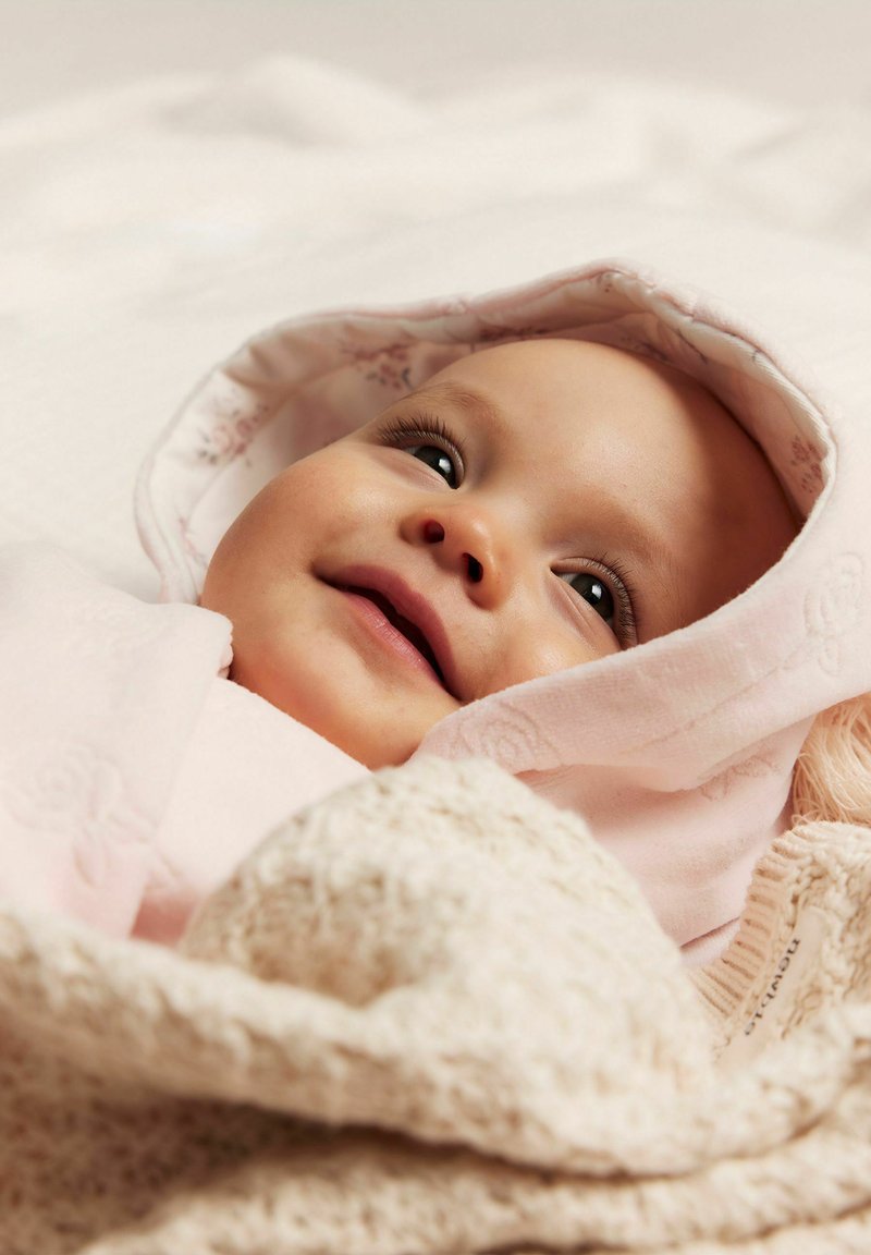 Lichtroze babydeken met bloemenkap, zachte textuur. Het gezicht van een baby met donkere ogen en een lichte glimlach, stevig gewikkeld in de stof.