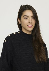 Pull en tricot noir avec un motif texturé, ornée de boutons dorés sur l'épaule et de détails en tricot torsadé sur les manches.