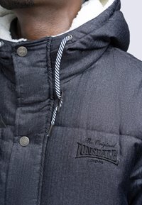 Svart vadderad jacka med fiskbensmönster, foder i päls, silverdetaljer och broderad logotyp. Justerbara dragsnören ingår.