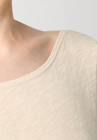 Licht beige top met een brede, afgeronde halslijn. De stof heeft een subtiele textuur met een lichte glans, met zachte, geblende vezels.