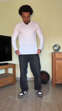 Camisa branca de Manga Longa combinada com jeans escuros e ténis preto e branco. De pé no interior, contra uma parede amarela clara.