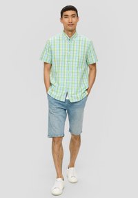 Camicia a maniche corte in plaid verde e blu, abbinata a pantaloni corti in denim chiaro. Modello che indossa sneakers bianche, in piedi con le mani in tasca.