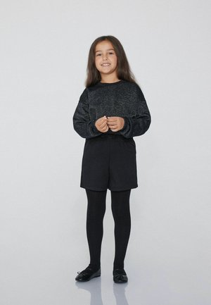 Camicia nera a maniche lunghe con un design texturizzato, abbinata a pantaloni corti neri e collant, completata da scarpe nere con dettagli a fiocco.