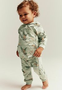 Grön tvådelad barnklädsel med en huvad zip-jacka och byxor. Tyget har ett naturmotiv med ekorrar och ormbunkar.