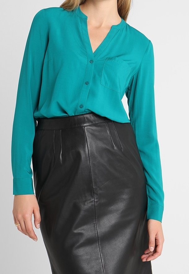 Femme portant un chemisier à manches longues turquoise boutonné, rentré dans une jupe en cuir noire taille haute, debout devant un fond uni.