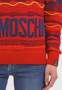 Červený pletený svetr s modrým nápisem "MOSCHINO", geometrickými vzory, žebrovanými manžetami a žlutými akcenty. Ruka spočívá na džínách.