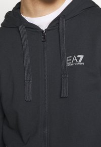 Šedá mikina na zip s texturovanou kapucí na šňůrku; má malé logo EA7 na hrudi v bílé barvě. Měkká tkanina, neformální design.