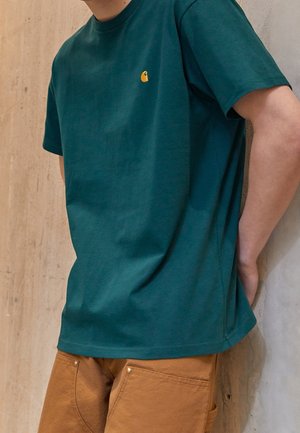 Person iført en teal kortærmet t-shirt med et gult logo og brune bukser, stående foran en beige struktureret væg.